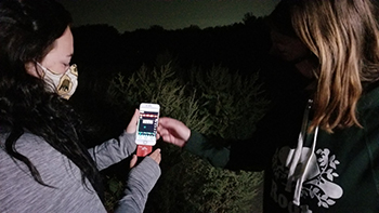 Two people standing out in a darkened area, look at graphics on their mobile phone, using the Echo Meter Touch Pro 2.
