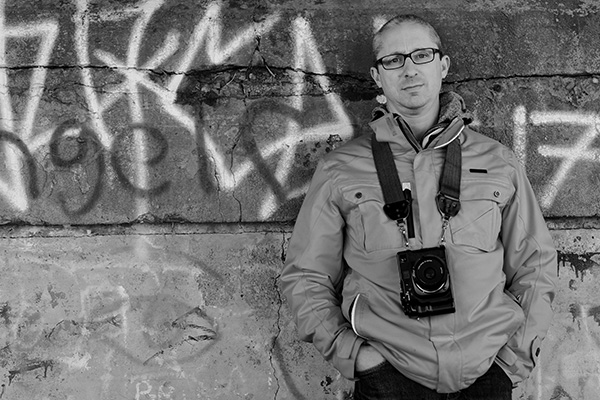 photo of man wearing a camera around his neck, leaning on graffiti covered wall