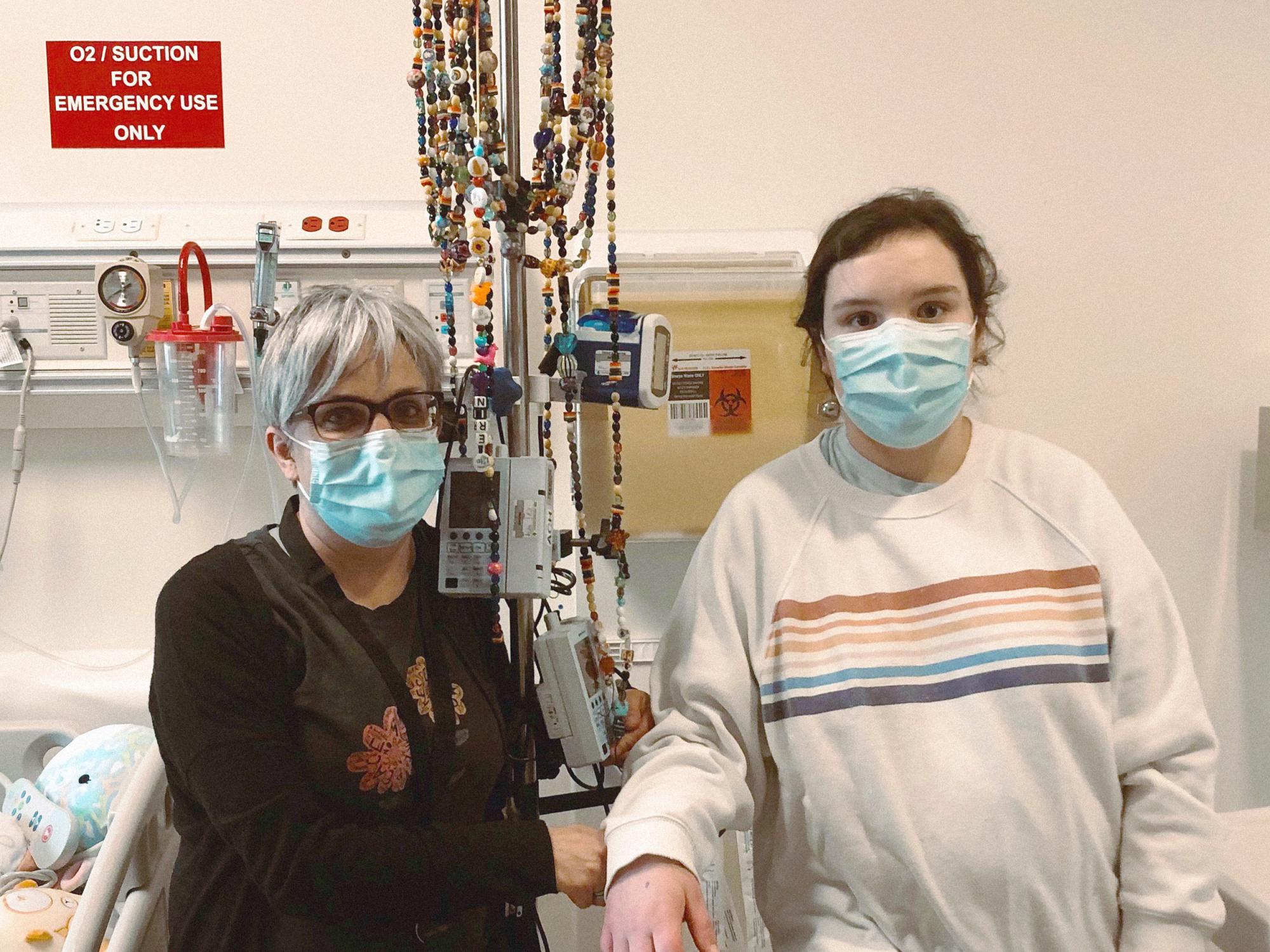 A woman and a teenaged girl wear surgical masks and pose in front of a intravenous stand in a hospital room.