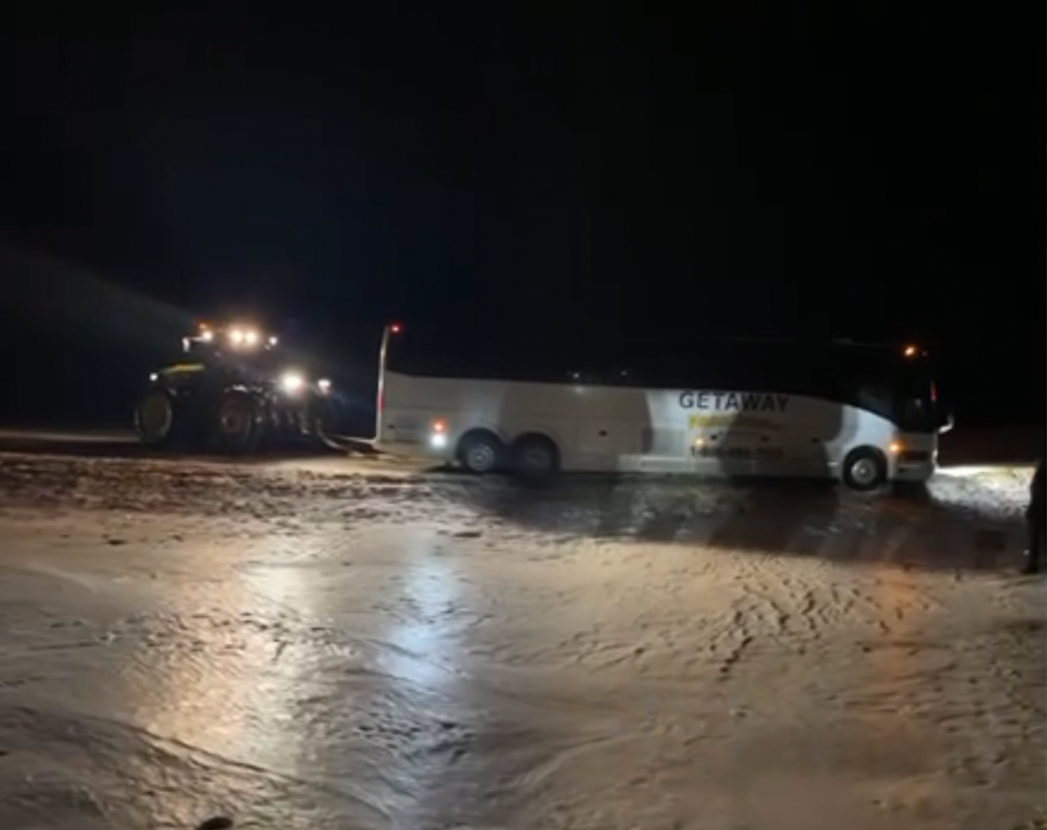 Tractor pulling bus out of snowbank on a dark road.