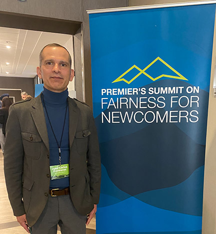 A man stands next to a sign that says "Premier's Summit on Fairness for Newcomers."