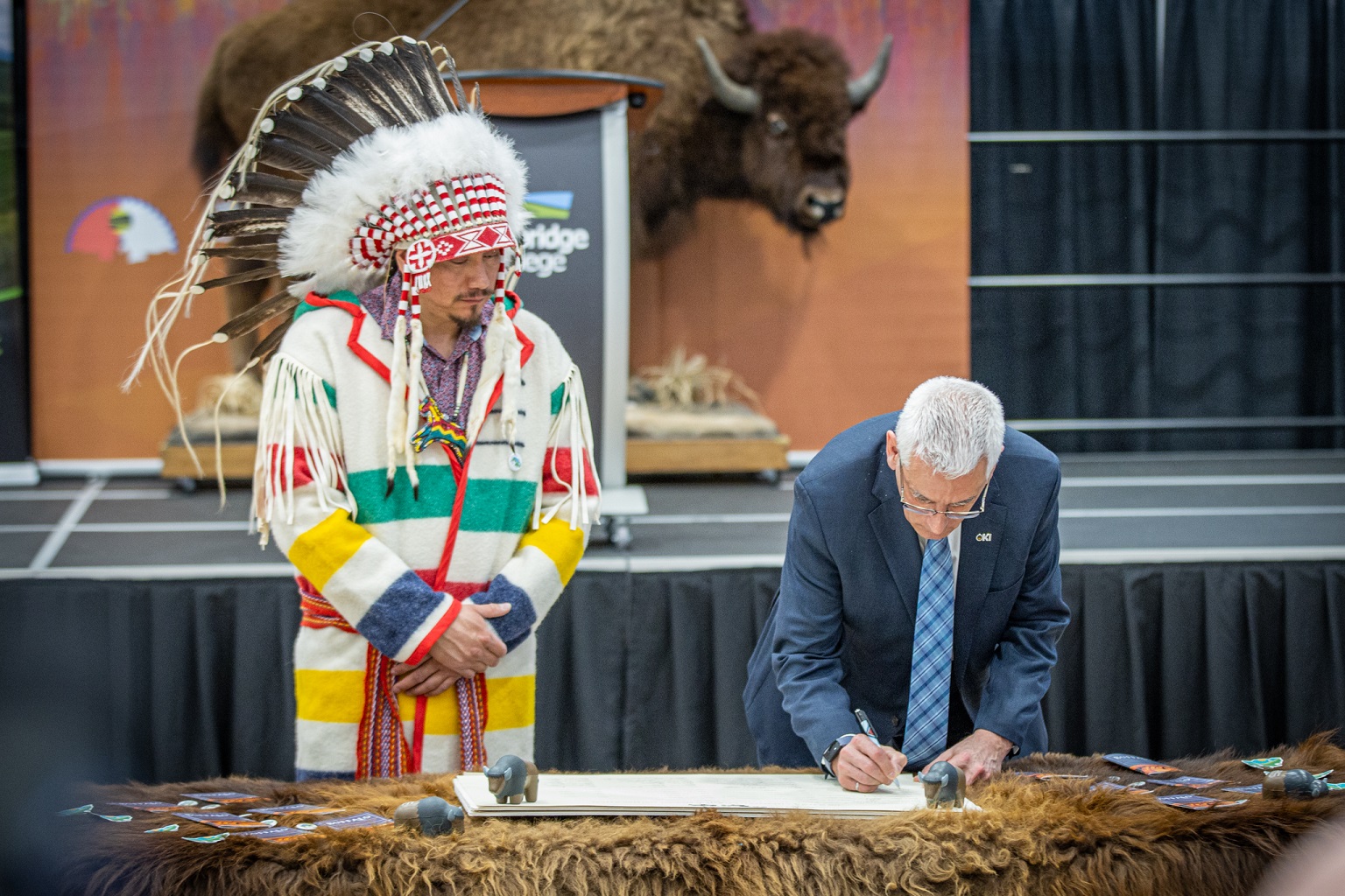 a man wearing a suit signs a pape while a man wearing traditional Indigenous clothing looks on