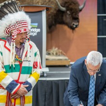 A man wearing a suit signs the Buffalo Treaty while a man wearing traditional Indigenous clothing looks on