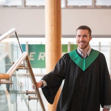 A man wearing a grad gown stands on the stairs and smiles for the camera